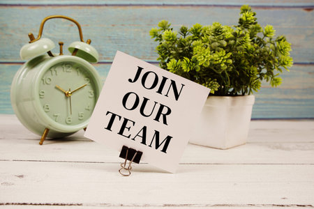 Join Our Team text written on paper card with alarm clock on wooden background
