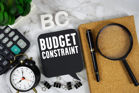 Budget Constraint text with Calculator, alarm clock and magnifying glass on marble background