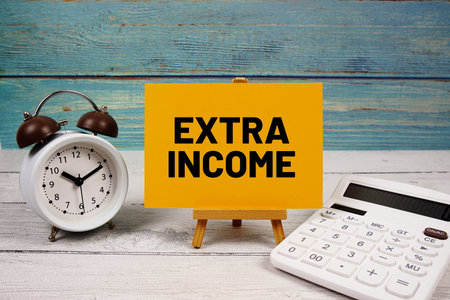 Extra Income text written on paper card with calculator and alarm clock on wooden backgroundの写真素材