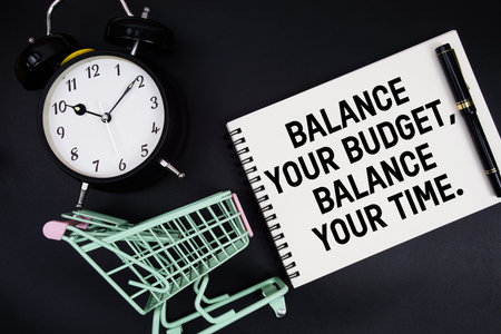 Balance Your Budget, Balance Your Time text written on notepad with alarm clock and trolley shopping...