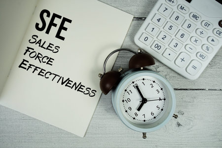 SFE Sales Force Effectiveness text on paper notebook page with alarm clock and calculator on wooden...