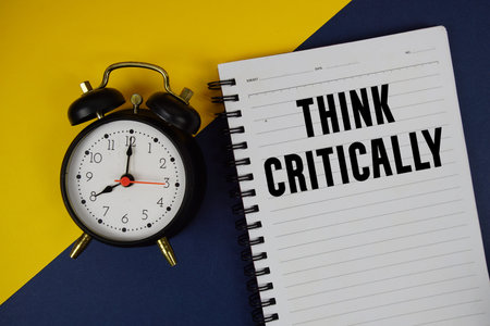 Think Critically text written on notepad with alarm clock on blue background