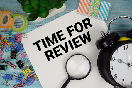 Time For Review text on a paper surrounded by office supplies, a magnifying glass, and a clock on a blue wooden surfaceの写真素材