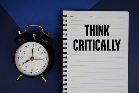 Think Critically text written on notepad with alarm clock on blue background
