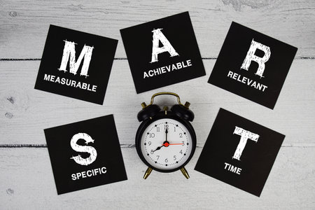 SMART Abbreviations of Specific, Measurable, Achievable, Relevant, Time text written on sticky note and alarm clock flat lay on wooden backgroundの写真素材
