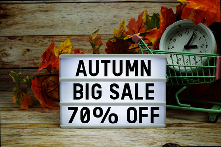 Autumn Big Sale 70% off text with trolley shopping cart and alarm clock decoration with maple leaf on wooden backgroundの写真素材
