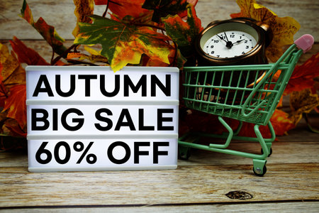 Autumn Big Sale 60% off text with trolley shopping cart and alarm clock decoration with maple leaf on wooden backgroundの写真素材