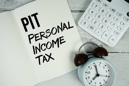 PIT Personal Income Tax text on paper notebook page with alarm clock and calculator on wooden backgroundの写真素材
