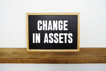 Blackboard with text CHANGE IN ASSETS on a wooden shelf, set against a white wall.