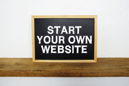 Start Your Own Website text written on blackboard with wooden shelf and concrete background