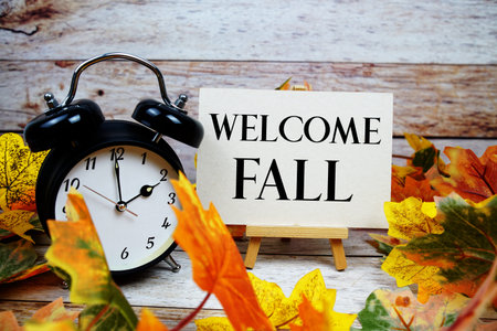 Welcome Fall text with alarm clock and maple leaf decoration on wooden background