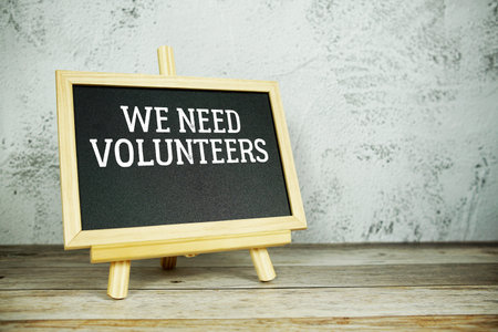 wooden easel displays a blackboard with the bold white text We Need Volunteers set against a rustic...