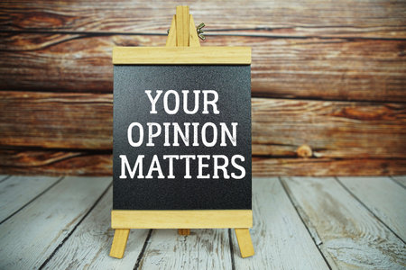 wooden easel holds a blackboard with the white text "Your Opinion Matters" set against a rustic wooden wall and light-colored wooden floorの写真素材