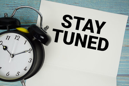 Notepad on a blue wooden table with the words Stay Tuned and alarm clock