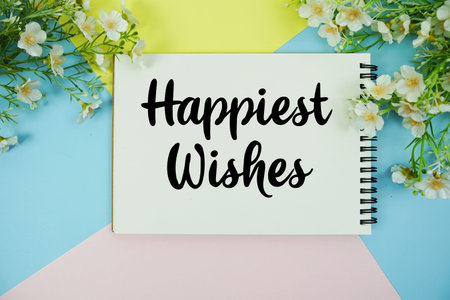 Happiest Wishes text written on notepad with flowers decorations flat lay on multicolor pastel background