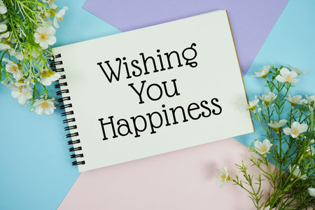 Wishing You Happiness text written on notepad with flowers decorations flat lay on multicolor pastel background