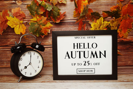Hello Autumn Sale 25% off text with alarm clock and maple leaf decoration on wooden background