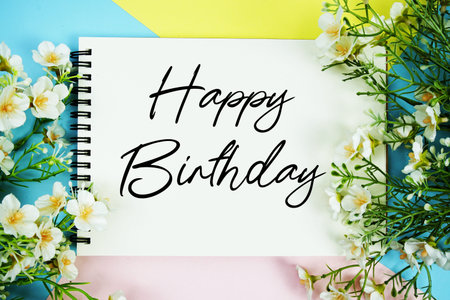 Happy Birthday text written on notepad with flowers decorations flat lay on multicolor pastel background