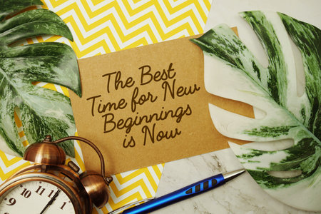 The Best Time for New Beginnings is Now text with alarm clock and monstera leave on marble background
