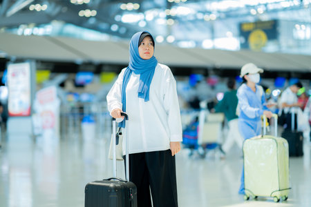 An Asian Muslim wearing a blue hijab is preparing for a vacation and she is at the airport. She is waiting for her friends, Muslim travelers.の写真素材