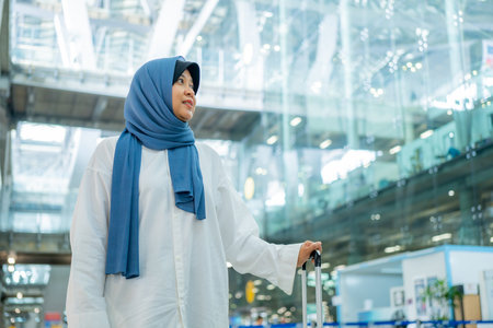 An Asian Muslim wearing a blue hijab is preparing for a vacation and she is at the airport. She is waiting for her friends, Muslim travelers.の写真素材