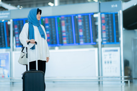 An Asian Muslim wearing a blue hijab is preparing for a vacation and she is at the airport, looking at the Flight Departure board.の写真素材