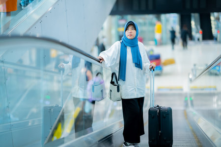 An Asian Muslim wearing a blue hijab is preparing for a vacation and she is at the airport. She is waiting for her friends, Muslim travelers.の写真素材