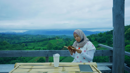beautiful Asian Muslim woman drinking hot coffee and reading the diary sitting on the chair mountain view in the morning.の写真素材