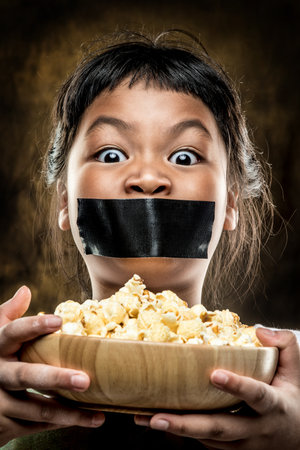 Asian girl with black tape on mouth and popcorn in wooden bowl.の写真素材
