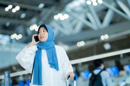 An Asian Muslim wearing a blue hijab is preparing for a vacation and she is at the airport. She is using her mobile phone to contact her friends and Muslim travelers.の写真素材
