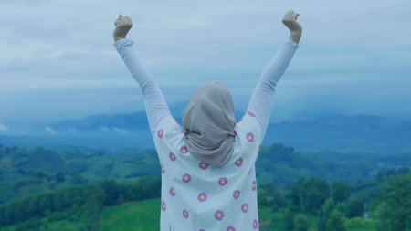 Asian Muslim woman standing at the top of the mountain looking at the view of nature, Enjoys the peace and quiet of the natural landscape with hills and trees in the backgroundの写真素材