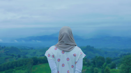 Asian Muslim woman standing at the top of the mountain looking at the view of nature, Enjoys the peace and quiet of the natural landscape with hills and trees in the backgroundの写真素材