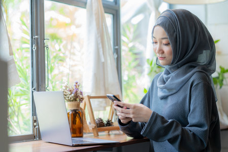 Modern Arab muslim girl in hijab using laptop and mobile phone at home, Cheerful girl sitting at desk and new order. Successful concept.の写真素材