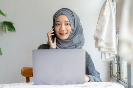Modern Arab muslim girl in hijab using laptop and mobile phone at home, Cheerful girl sitting at desk and new order. Successful concept.の写真素材