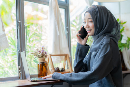 Modern Arab muslim girl in hijab using laptop and mobile phone at home, Cheerful girl sitting at desk and new order. Successful concept.の写真素材