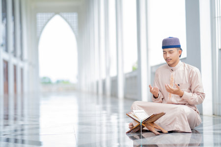 A little Asian Muslim man is sitting and reading the Quran. The peace in the mosque makes it an energetic atmosphere of faith, with copy space.の写真素材