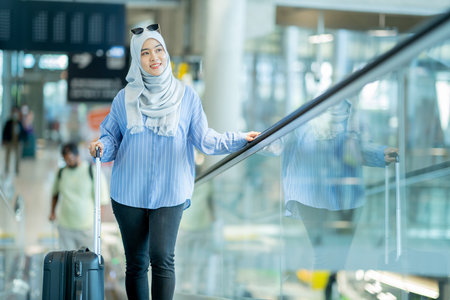 An Asian Muslim wearing a blue hijab is preparing for a vacation and she is at the international airport. She is waiting for her friends, Muslim travelers.の写真素材