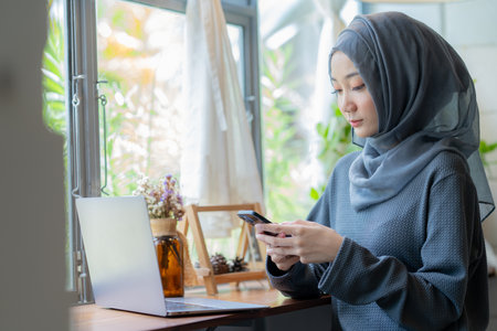 Modern Arab muslim girl in hijab using a laptop computer and mobile phone at home, Cheerful girl sitting at the desk and new order. Successful concept.の写真素材