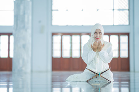 The image of an Asian Muslim woman in the Islamic religion in hijab in cream color. reading the Quran and having a happy smiling face Staying in a beautiful mosqueの写真素材