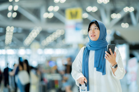 An Asian Muslim wearing a blue hijab is preparing for a vacation and she is at the airport. She is using her mobile phone to contact her friends and Muslim travelers.の写真素材