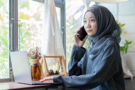 Modern Arab muslim girl in hijab using a laptop computer and mobile phone at home, Cheerful girl sitting at the desk and new order. Successful concept.の写真素材