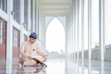 An Asian Muslim man is sitting and reading the Quran. The peace in the mosque makes it an energetic atmosphere of faith, with copy space.の写真素材