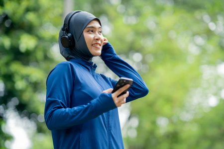 Asian cute muslim woman wearing sportswear and a blue hijab Working out and wearing earphones listening to music on her phone outdoors at the park in the morning with a fresh feeliの写真素材