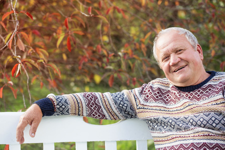 elderly man resting on white wooden bench in autumn parkの写真素材