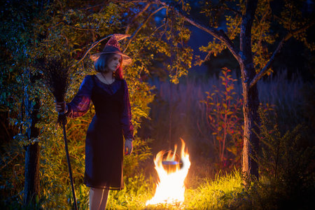 young witch by  fire in night forest makes in cauldron magic potion