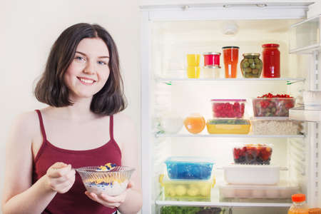 girl is having breakfast with cereals with milk and berries by open refrigeratorの写真素材