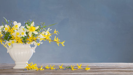 white and yellow spring flowers in vase on wooden table on blue background
