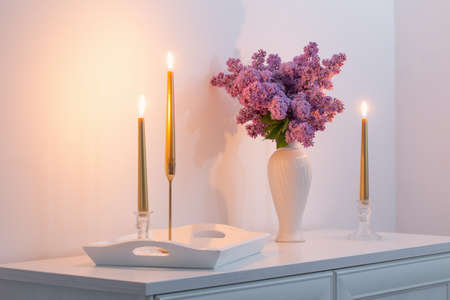 lilac flowers in white vase and golden candles on background white wallの写真素材