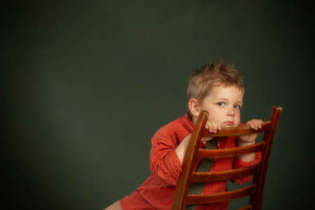 little sad boy in red shirt on old chair on dark green backgroundの写真素材