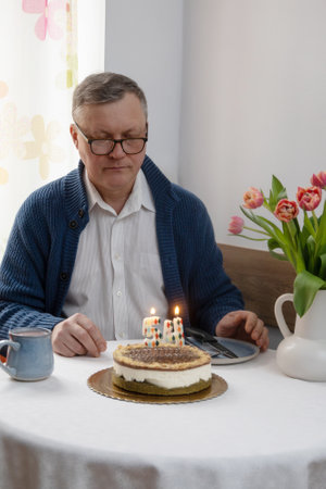 man looking at burning candles on his birthday cake alone in the kitchenの写真素材